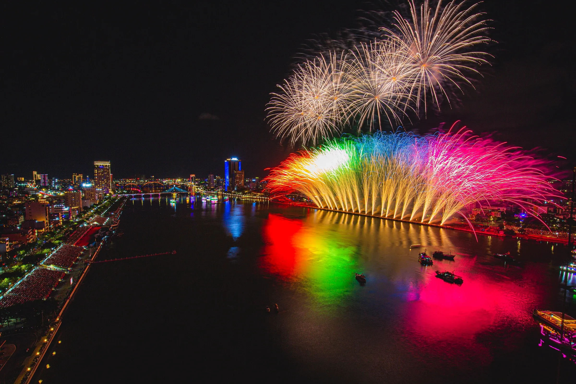 Fireworks bursting over the Han River from a launch barge, seen from above with the city lights beyond