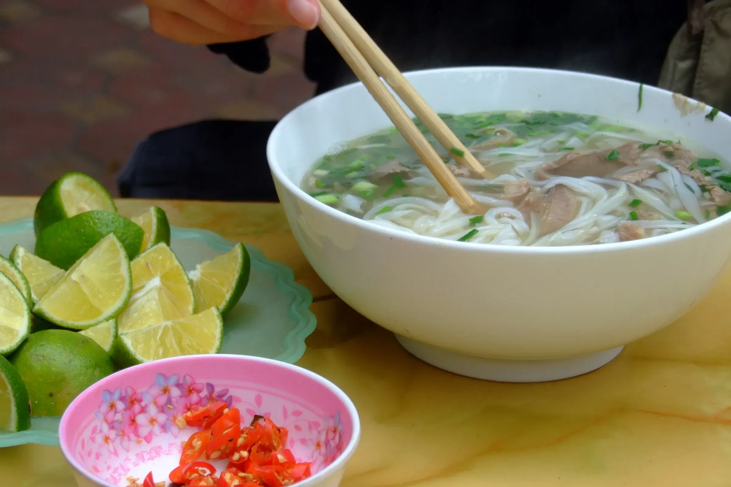 A bowl of beef pho with chopsticks, lime wedges and chilli on the side, Hanoi