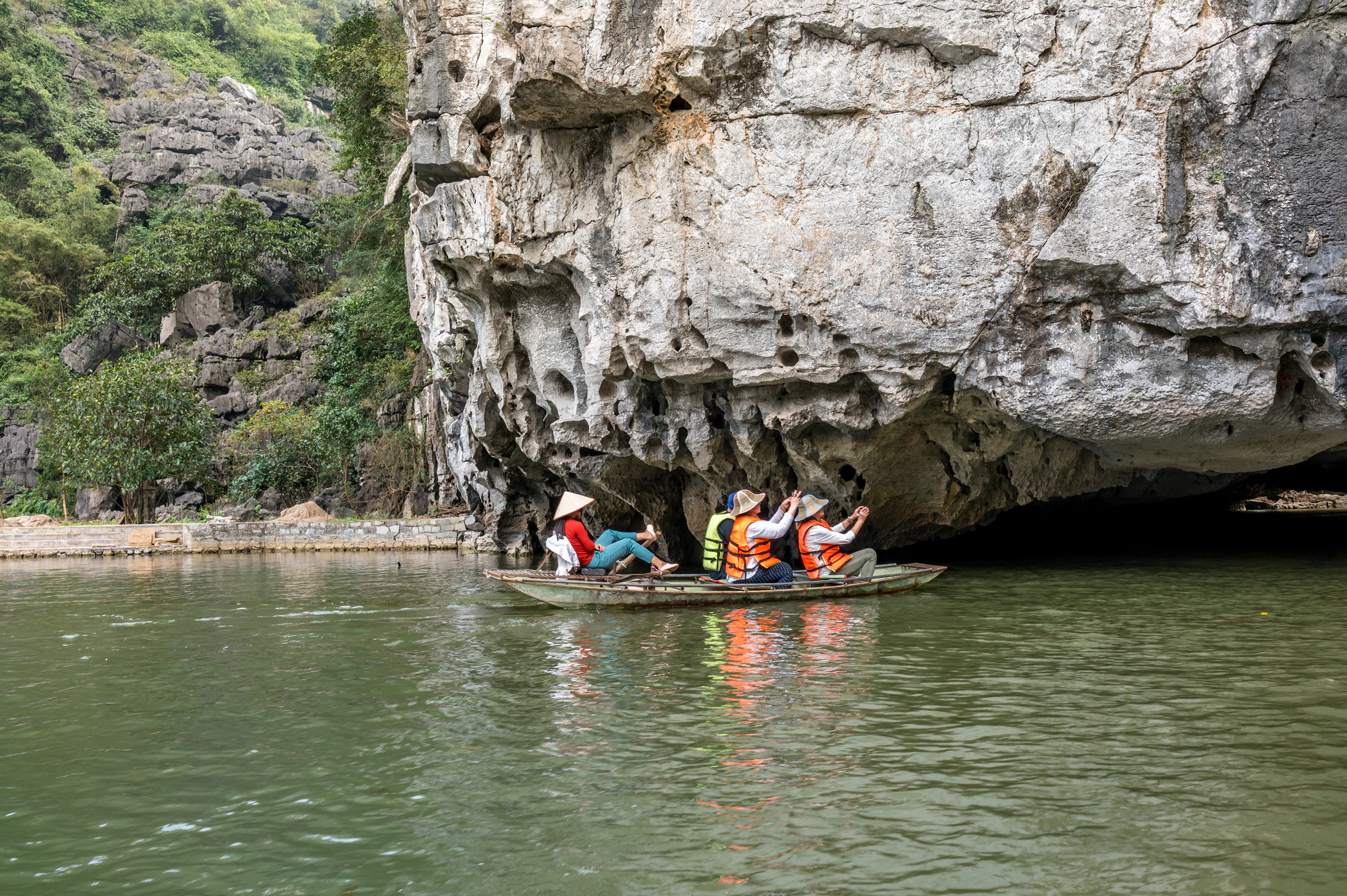 A boat of visitors in life jackets under a limestone overhang on a Ninh Binh river