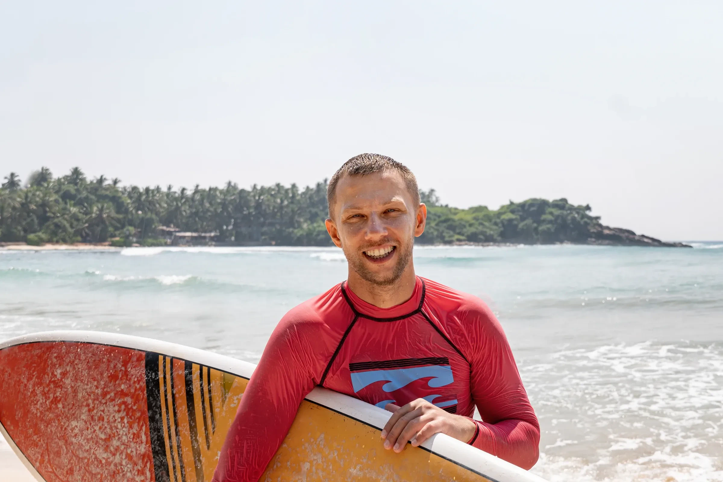 A smiling surfer in a rash vest holding a board on a Sri Lankan south coast beach