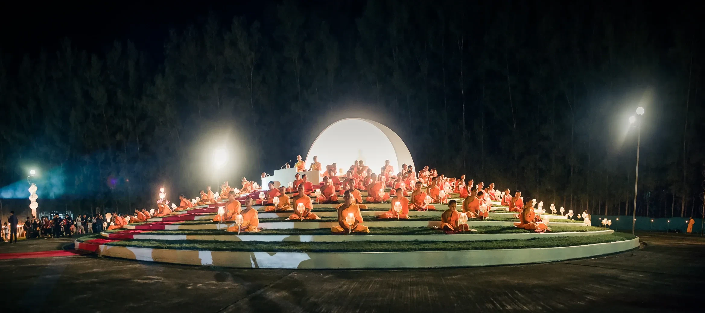 Monks sitting in rows on a lit platform beside a large white lantern during a Yi Peng ceremony in Chiang Mai