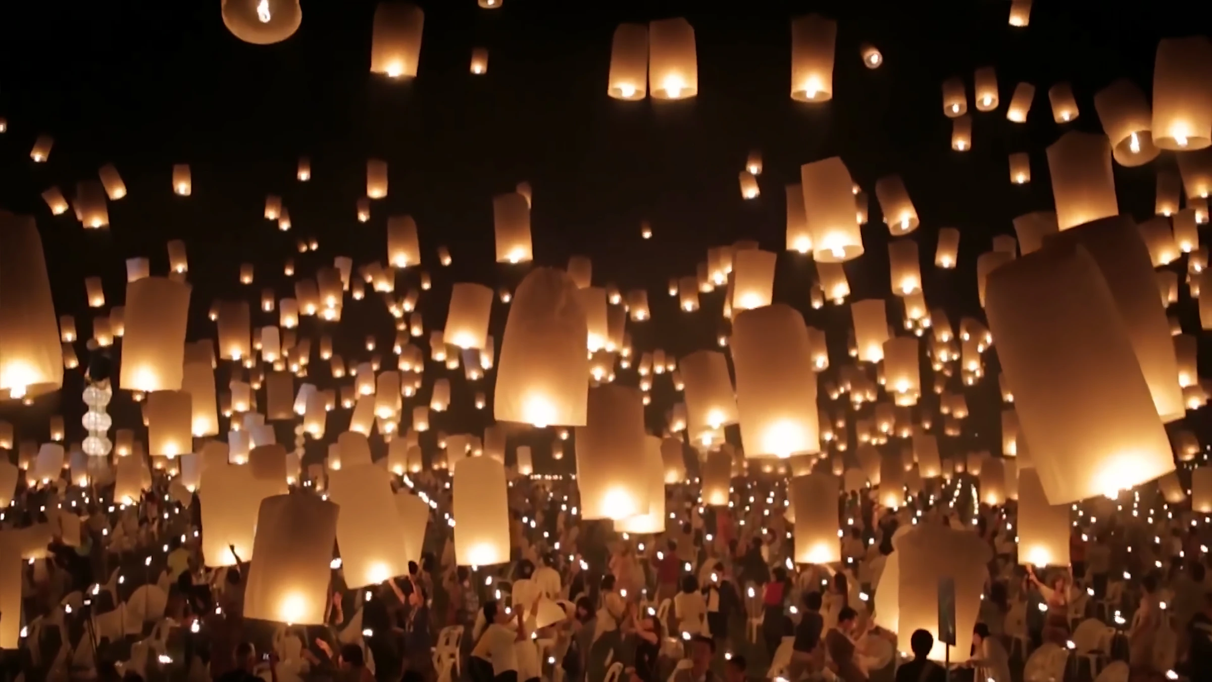 Hundreds of glowing sky lanterns rising above a crowd of people dressed in white at night