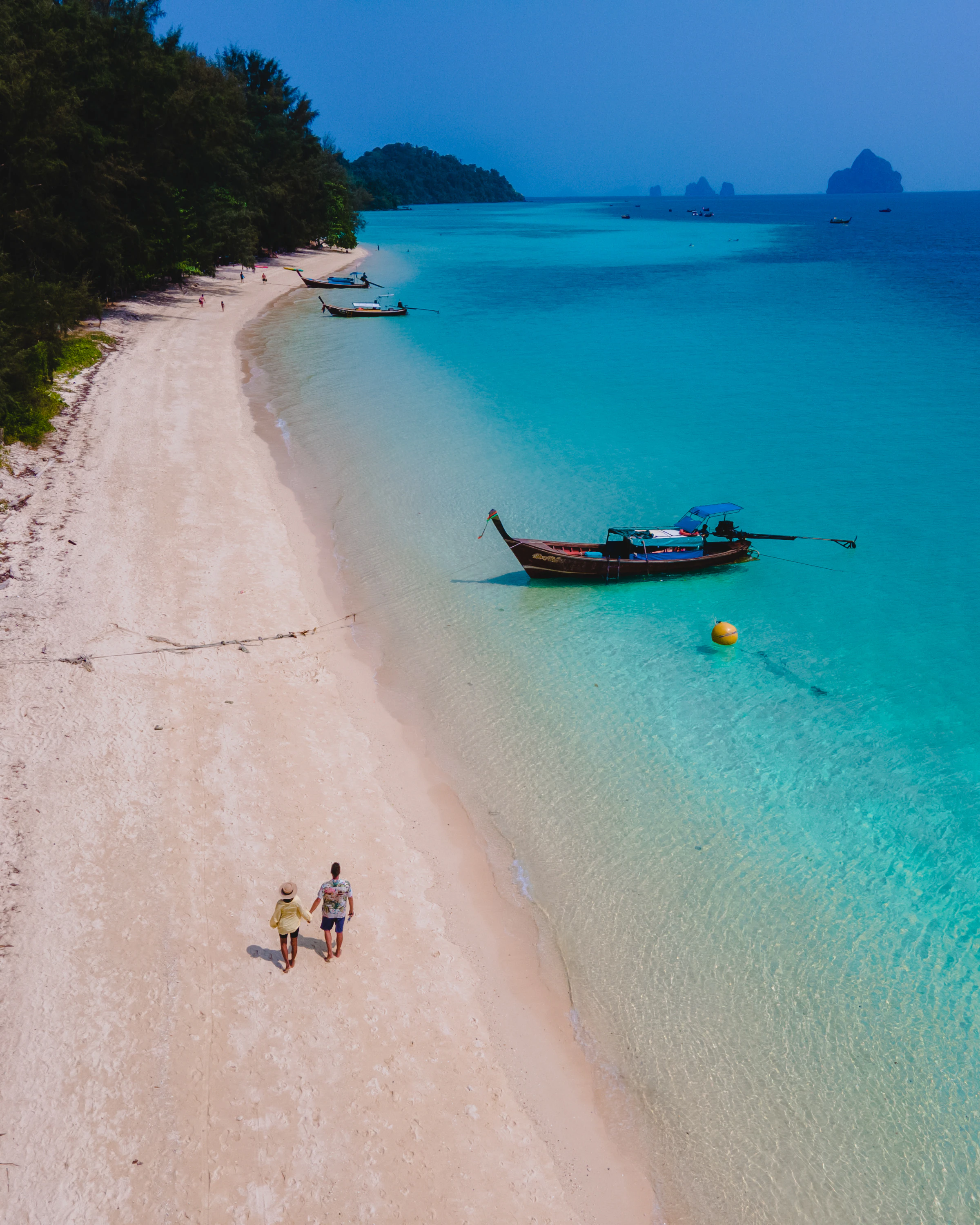 Aerial view of a couple walking along the white sand beach of Koh Kradan Island, Thailand, with a longtail boat in turquoise water