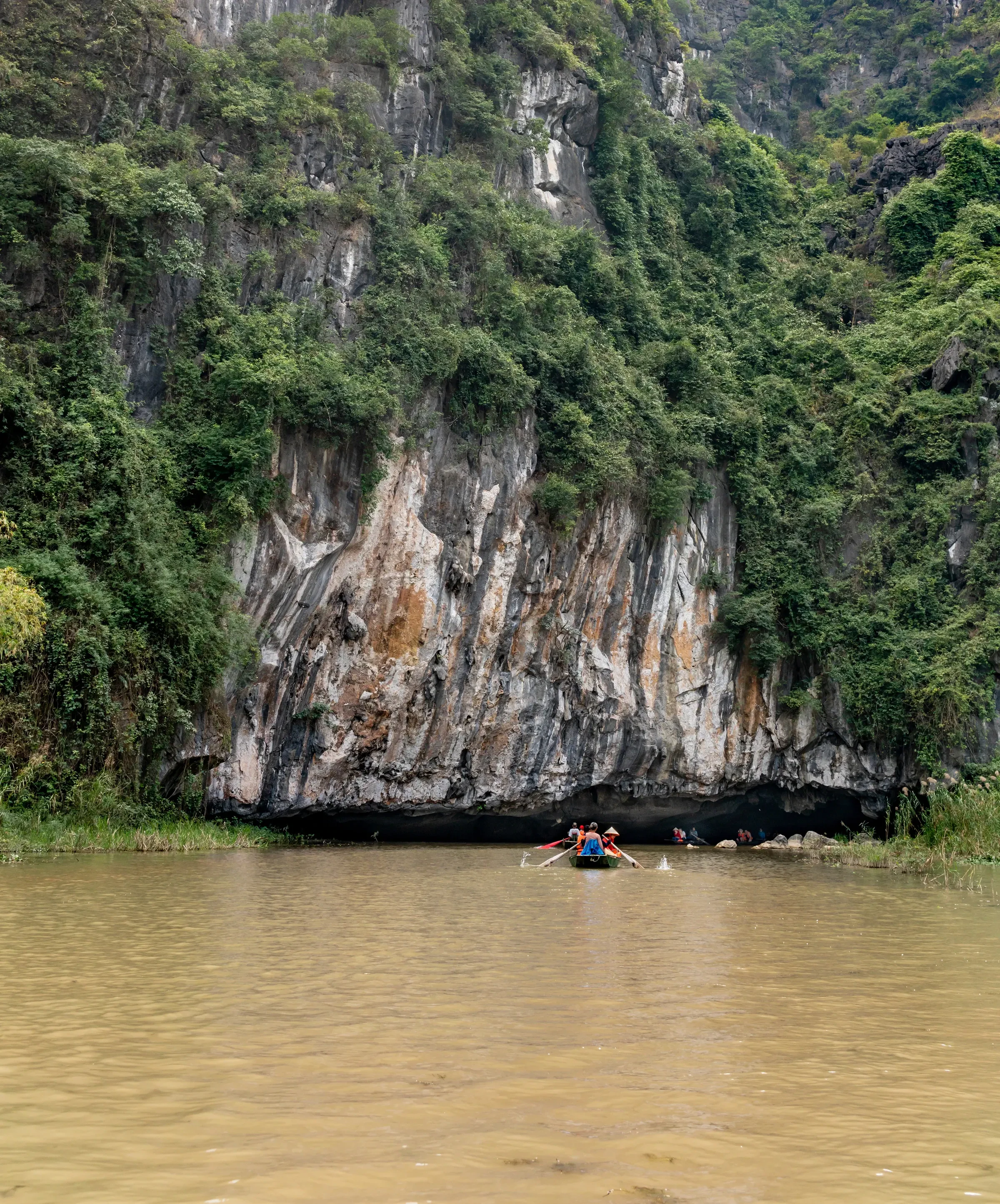 Tourists in a small rowing boat passing beneath a limestone cliff on a river in Vietnam