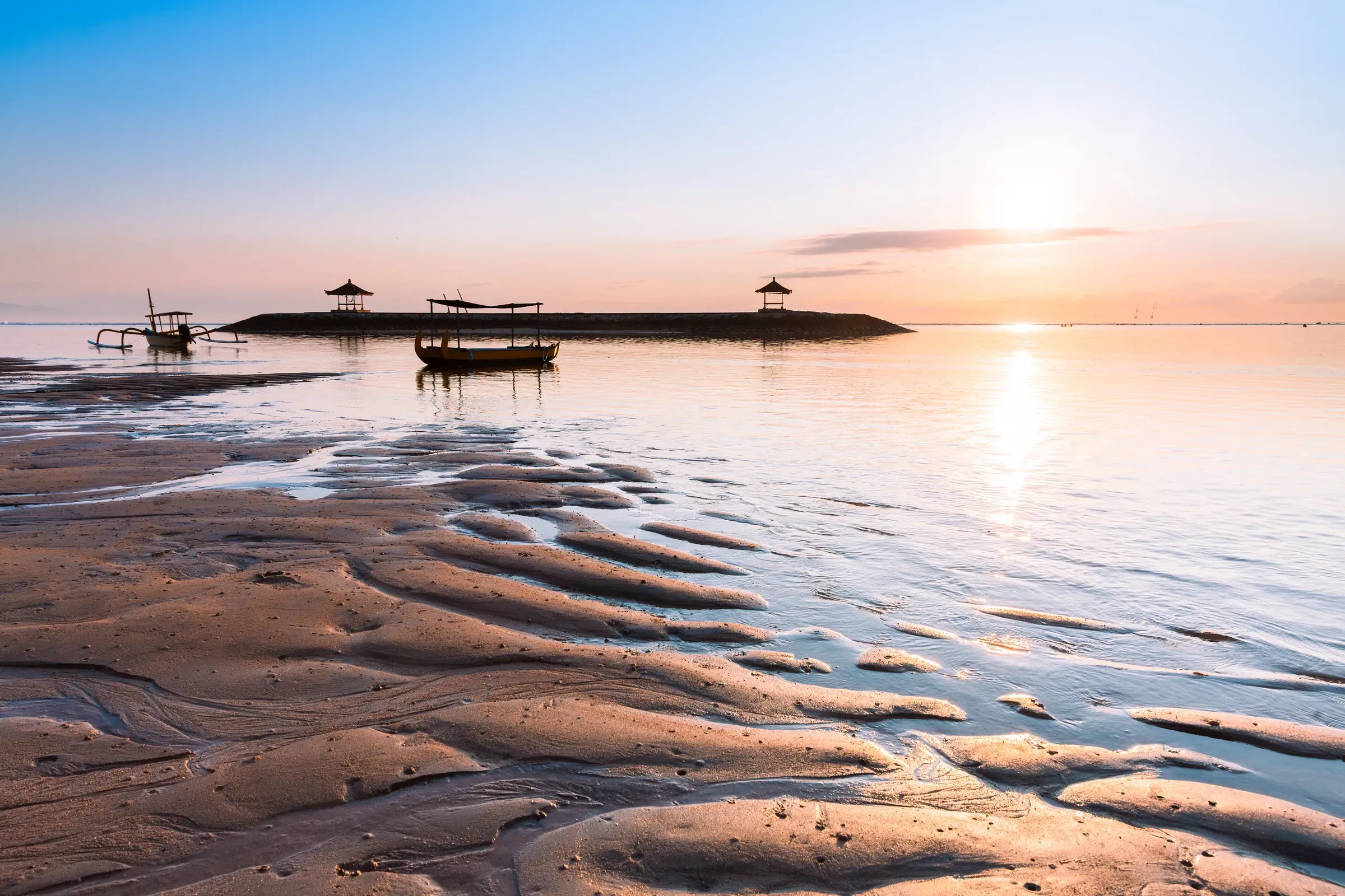 Sanur beach at low tide at sunrise, with a jukung boat and beach pavilions in the distance