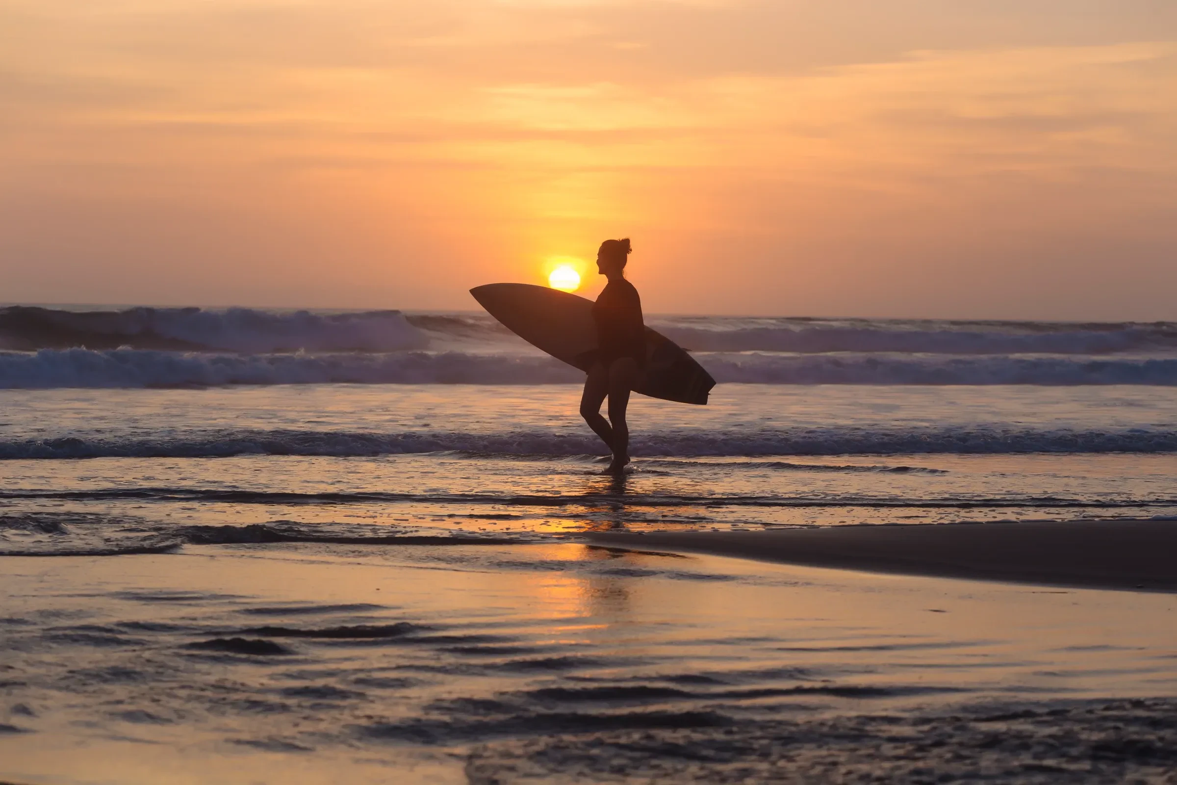 A surfer walking the shore at sunset with a board under one arm