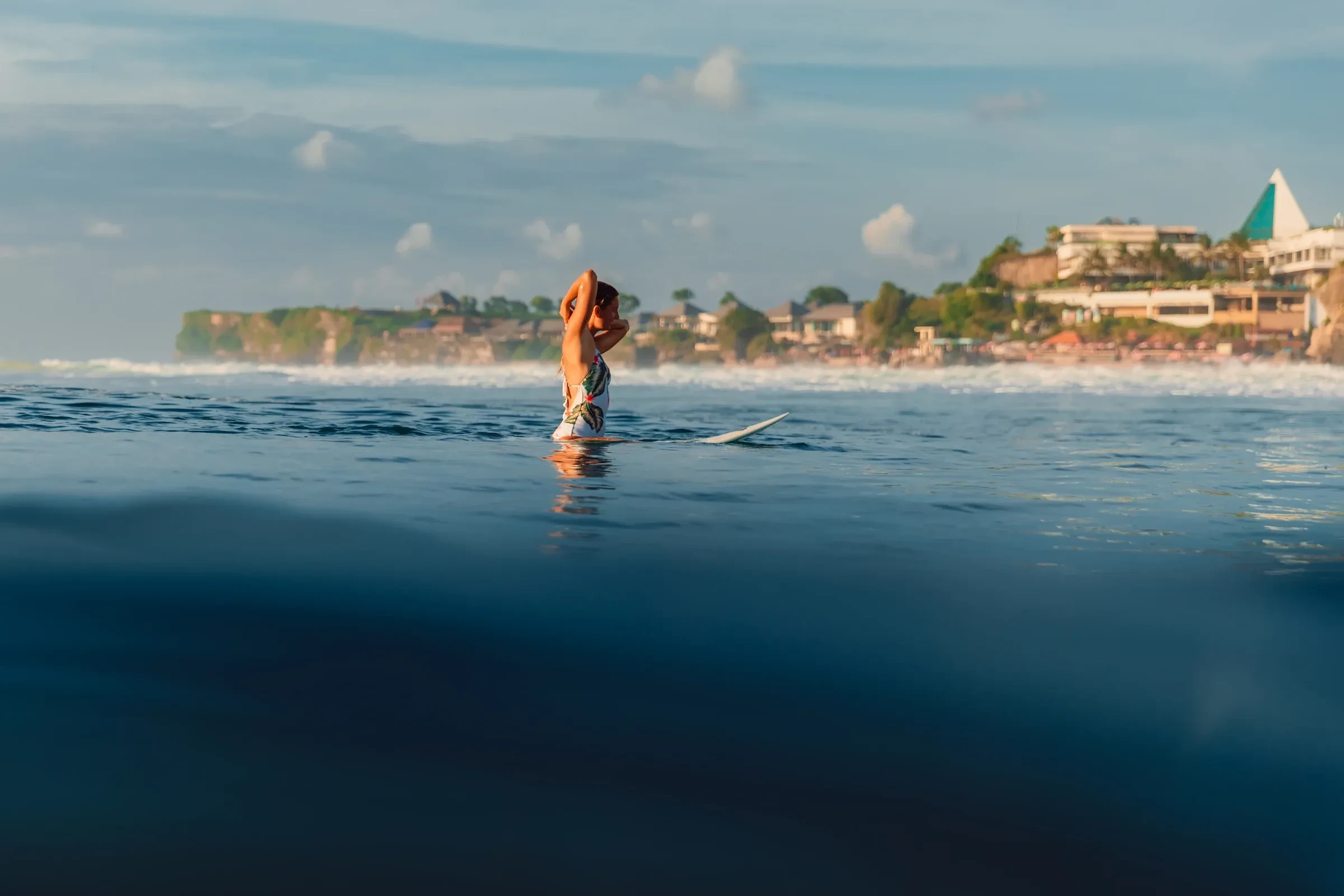 A surfer sitting on her board in calm water off a Bali beach