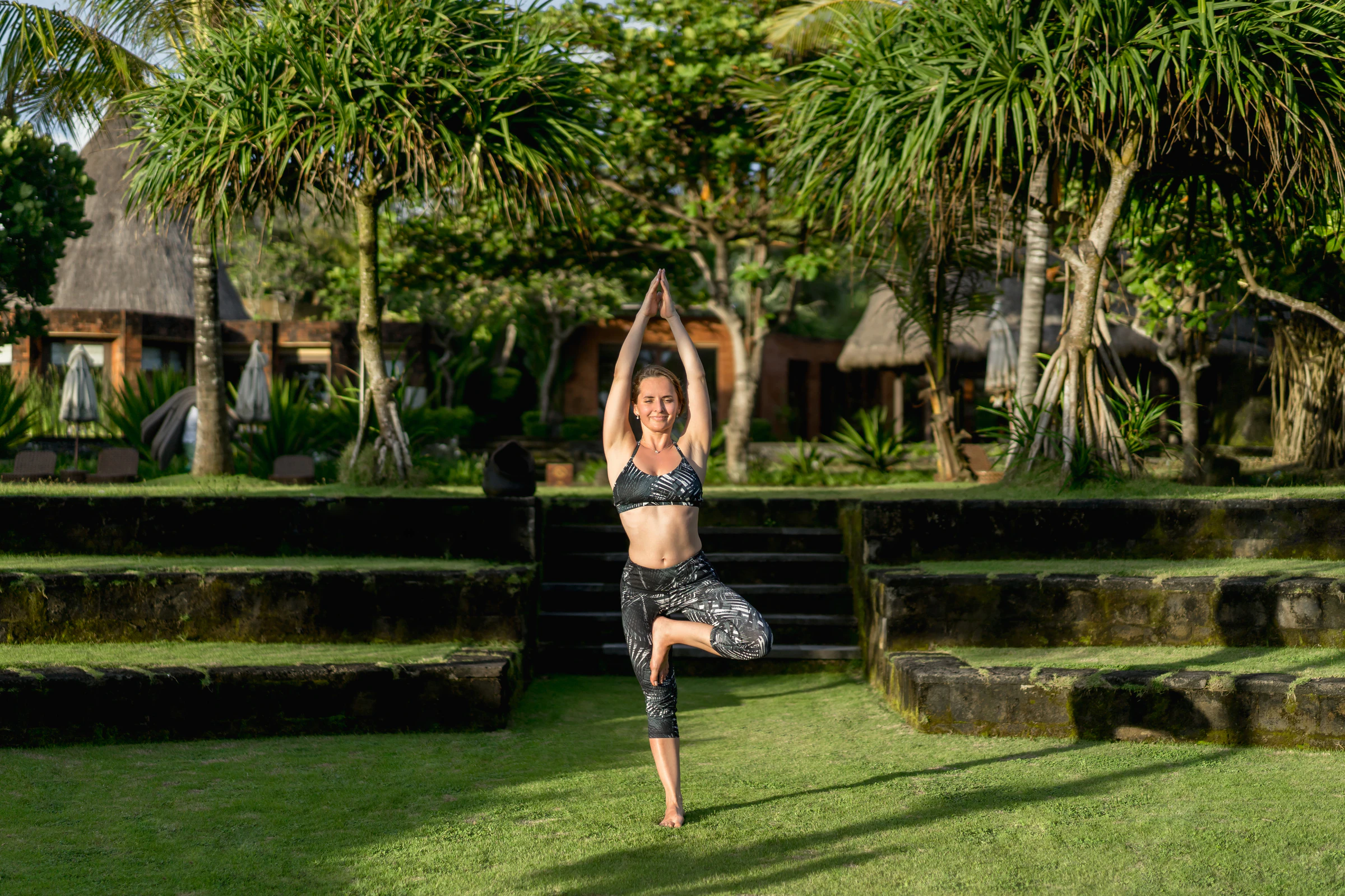 A woman in tree pose on a lawn beside tropical trees