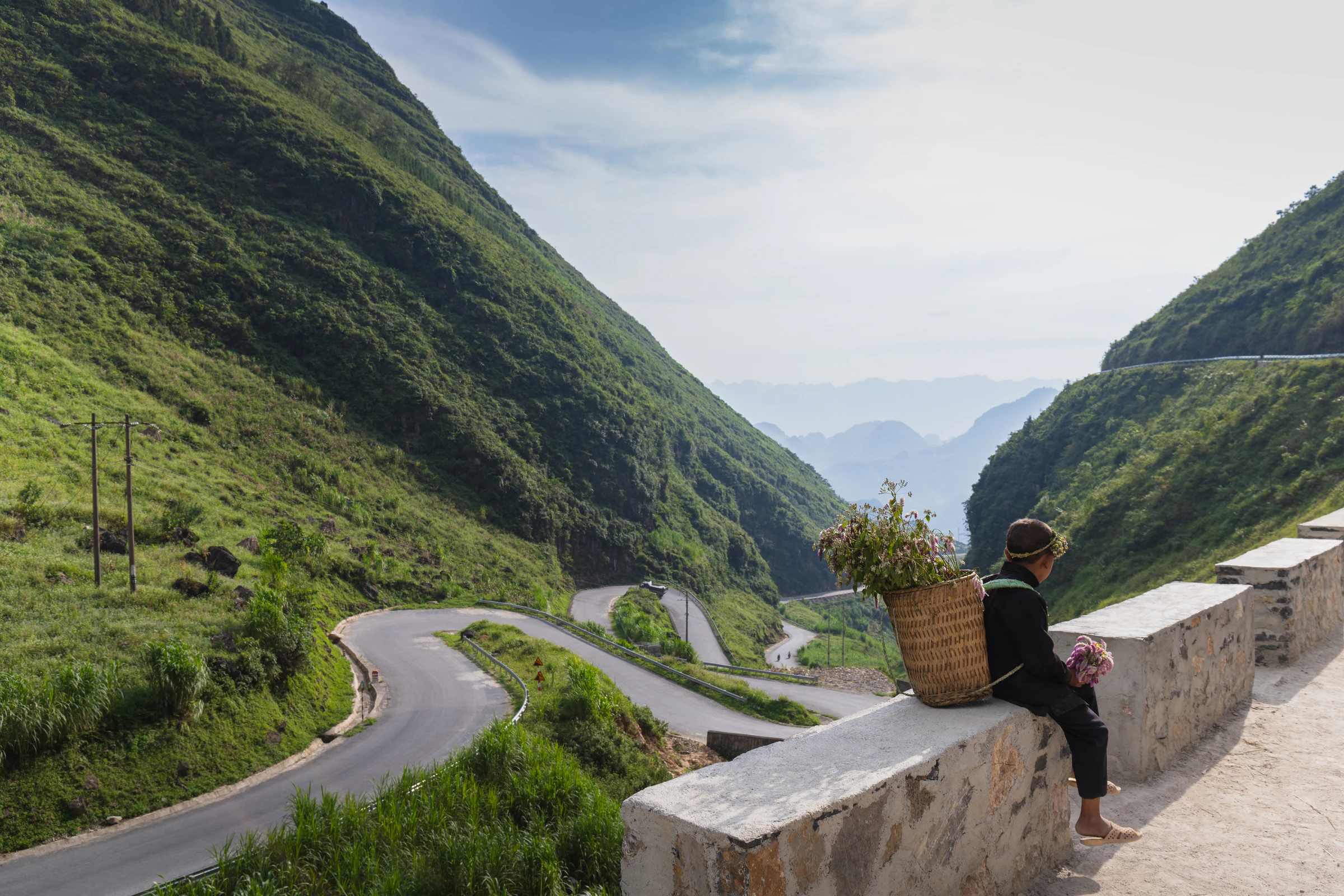 A road winding down a green hillside with a local woman resting on the wall