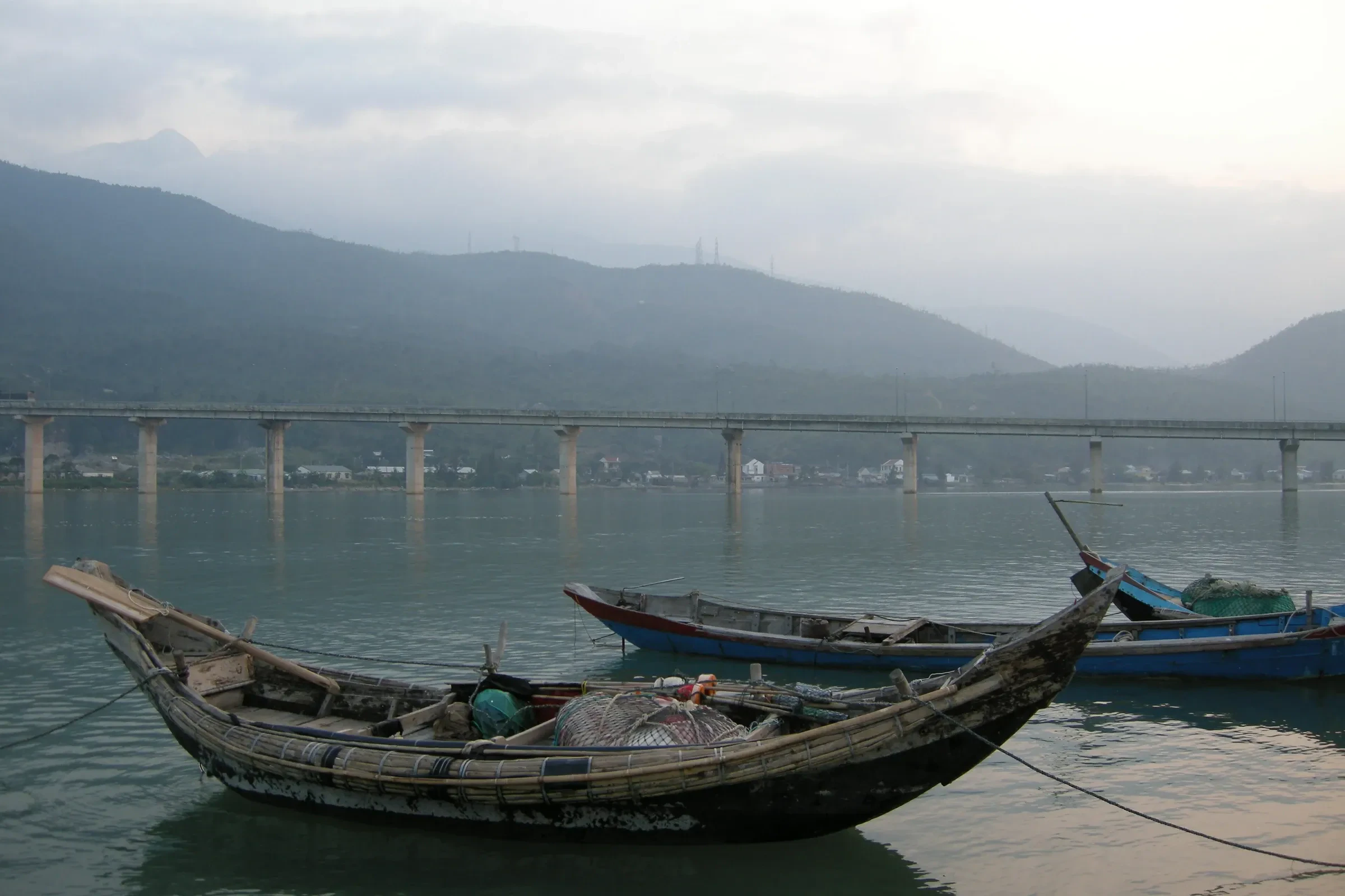 Wooden fishing boats on Lang Co lagoon, with the coastal bridge and hills behind