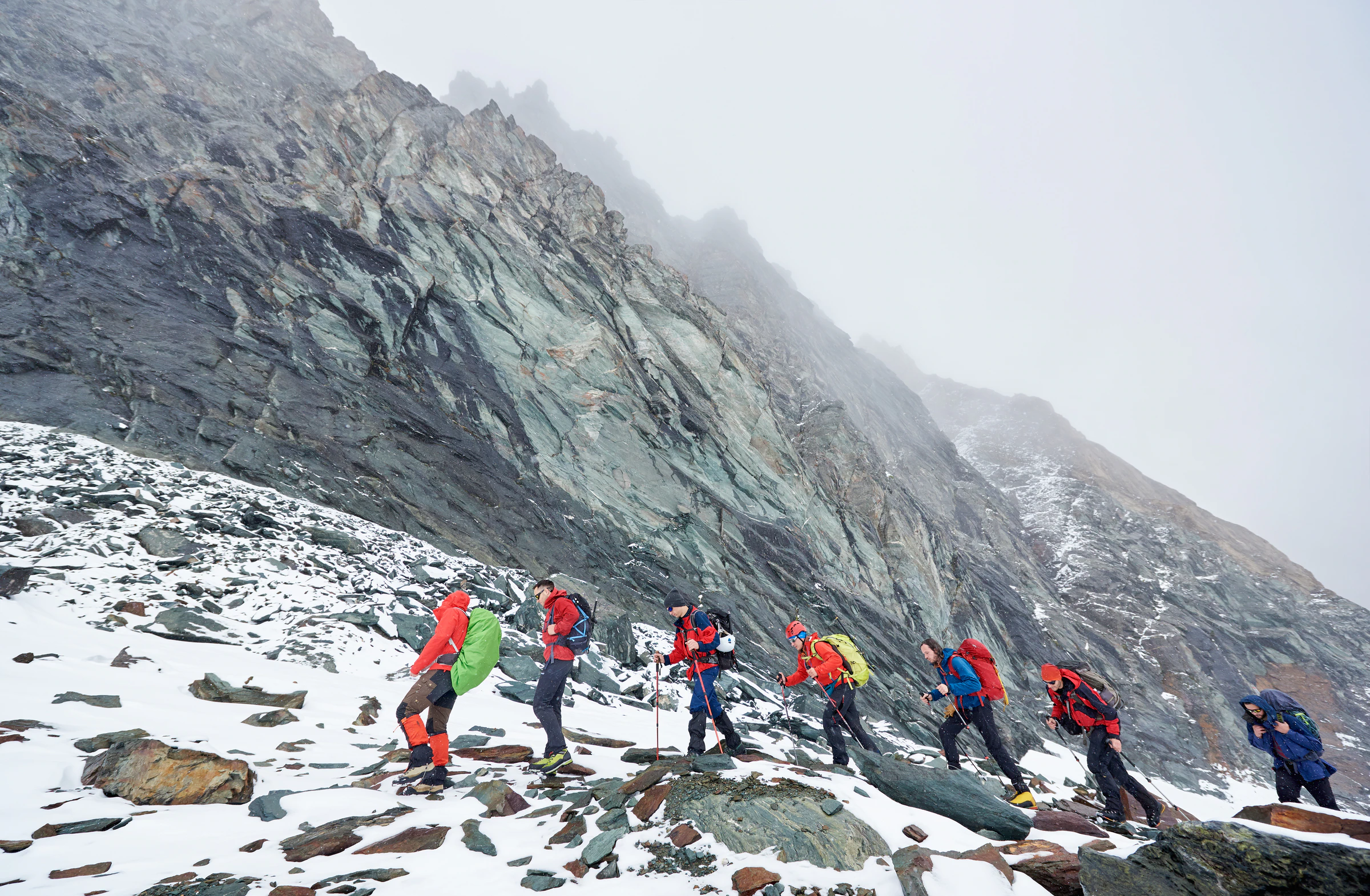 Trekkers in bright jackets walking across a snowy slope below a rocky ridge