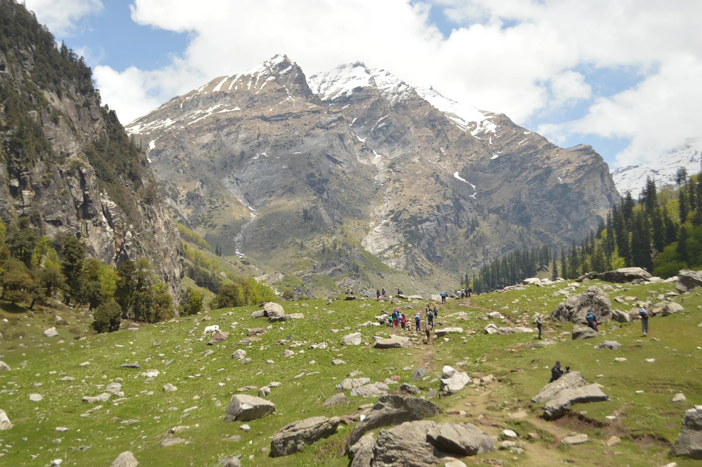 Trekkers crossing a green meadow below snow-streaked peaks on the way to Hampta Pass