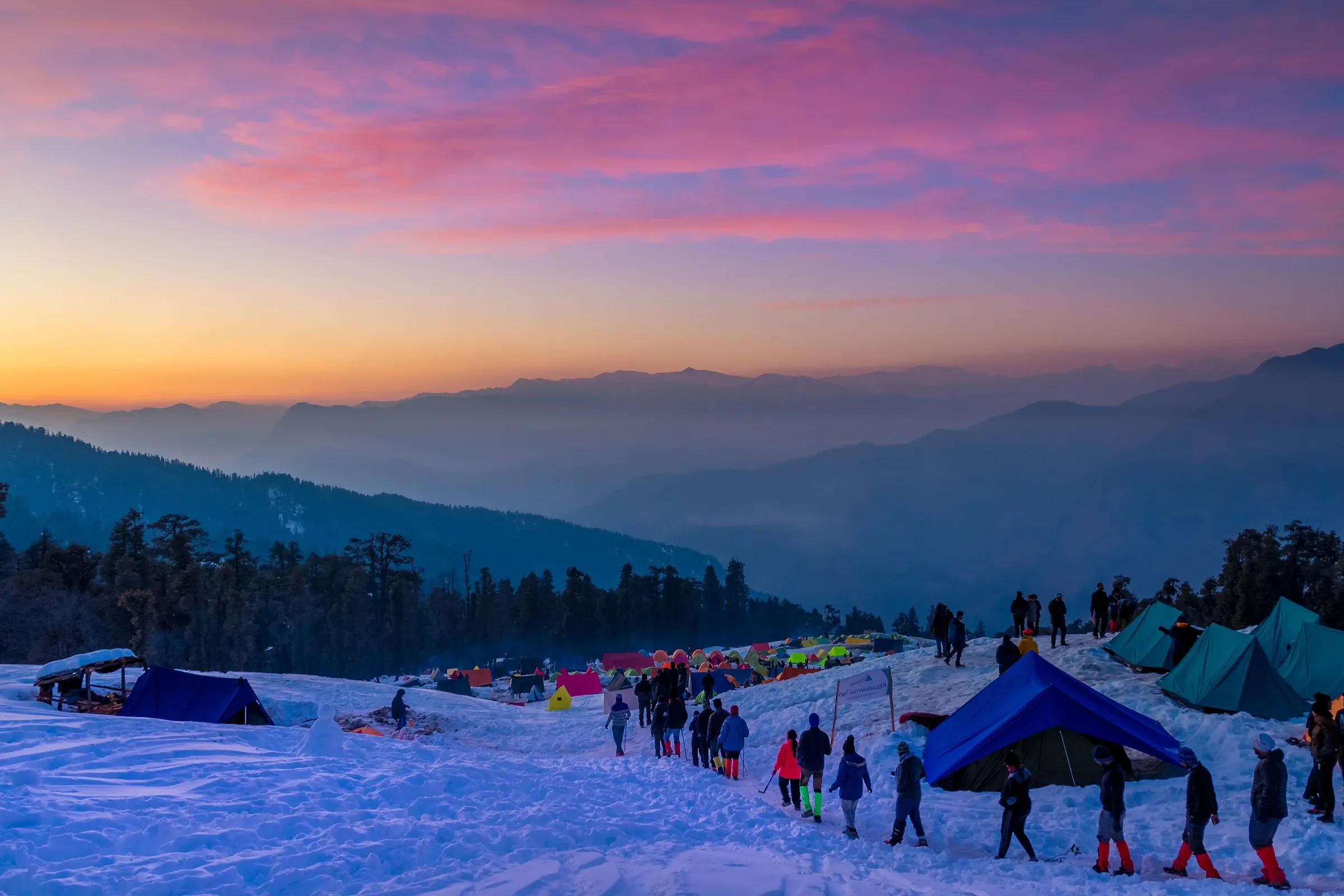 Trekkers and orange tents in a snow camp under a pink dusk sky