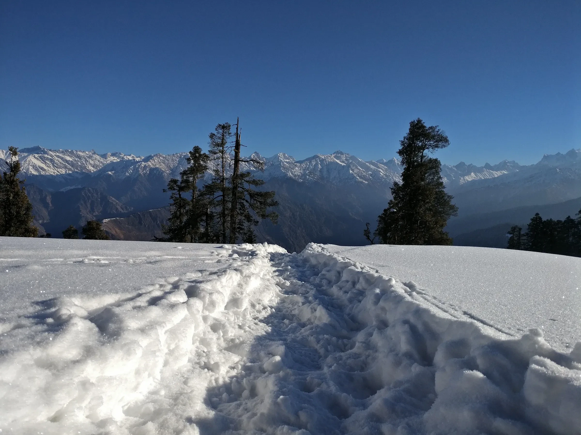 A snowy trail along a ridge with the Himalayan range beyond on the Kedarkantha trek