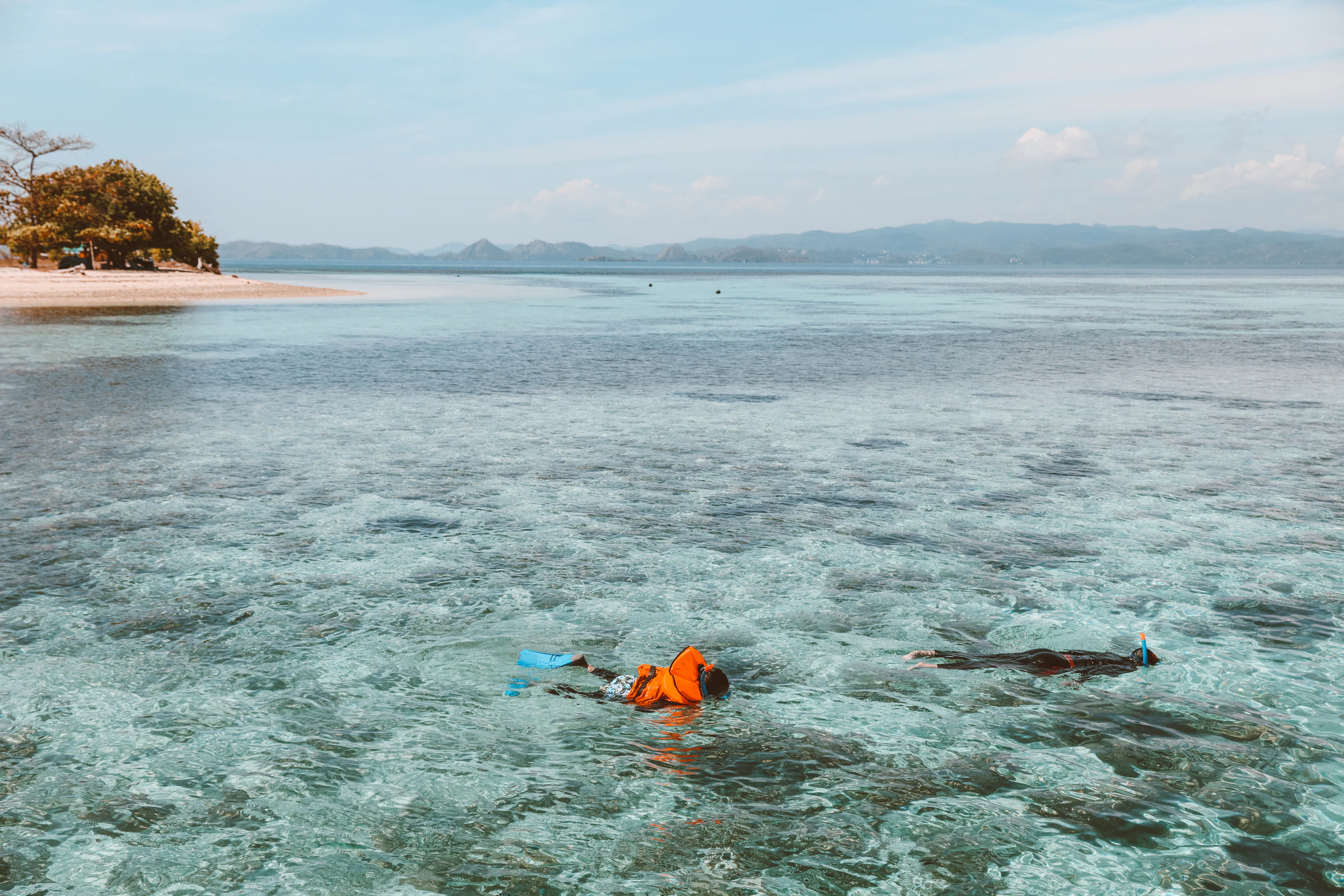 A snorkeller in an orange life jacket over clear shallows off a small island beach