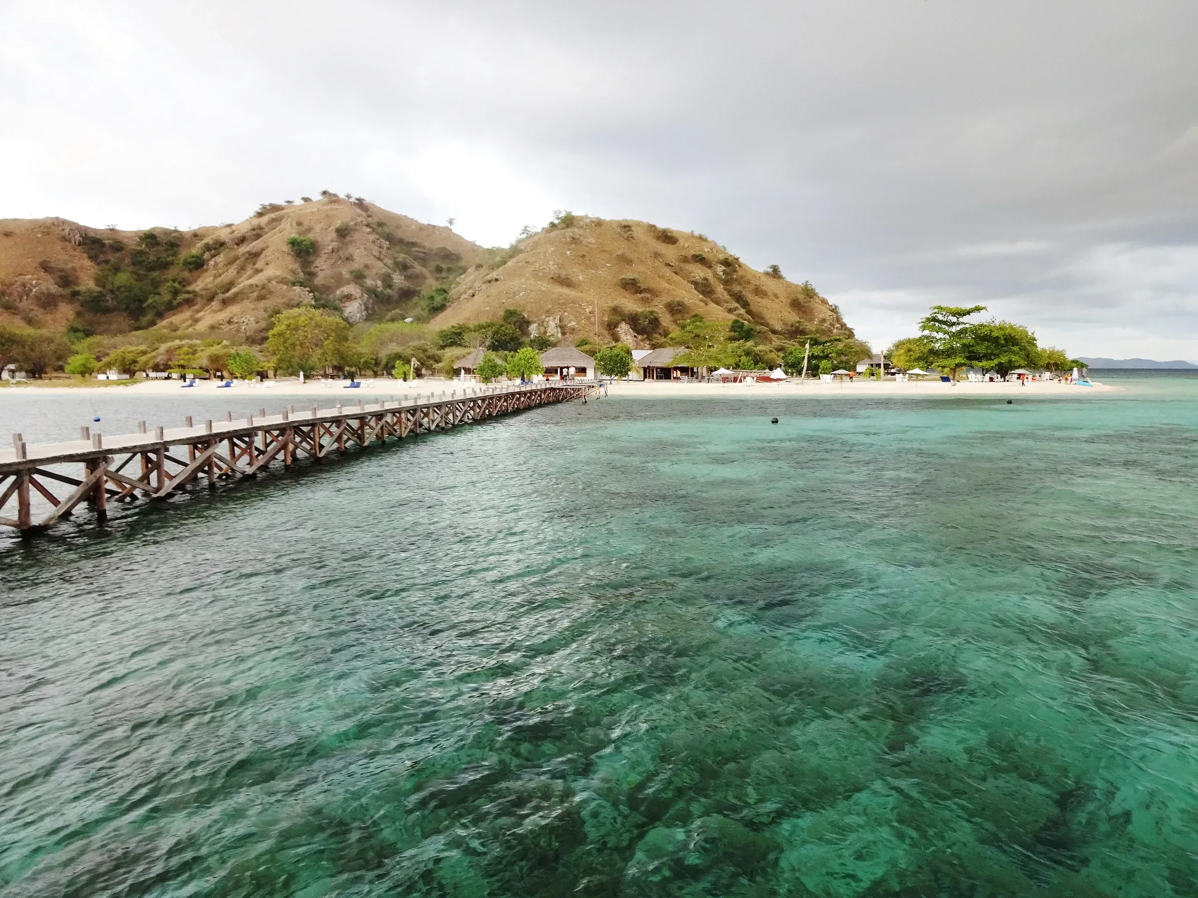 A wooden jetty and beach on Kanawa island with dry hills behind and clear green water