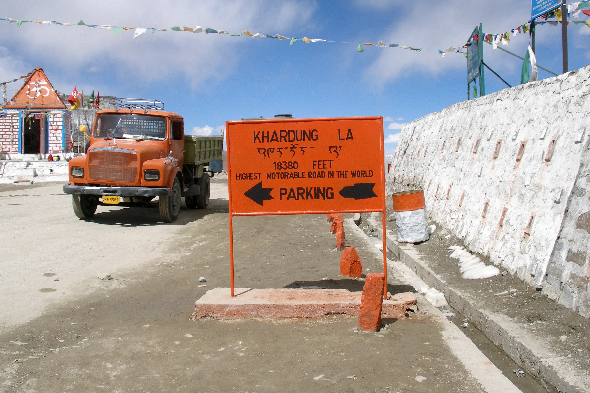 The orange Khardung La summit sign and prayer flags beside the road, with a truck parked at the top