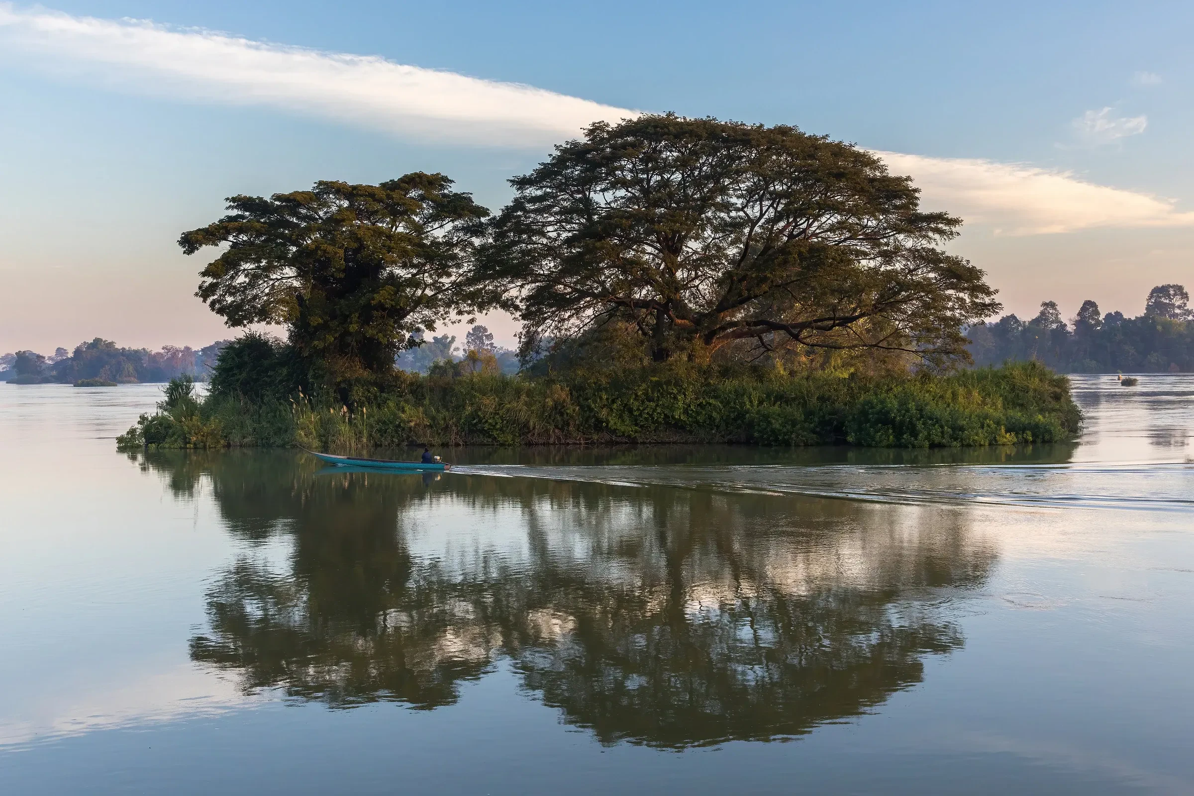 A tree-covered island reflected in the Mekong near Don Det at sunrise, with a small boat passing