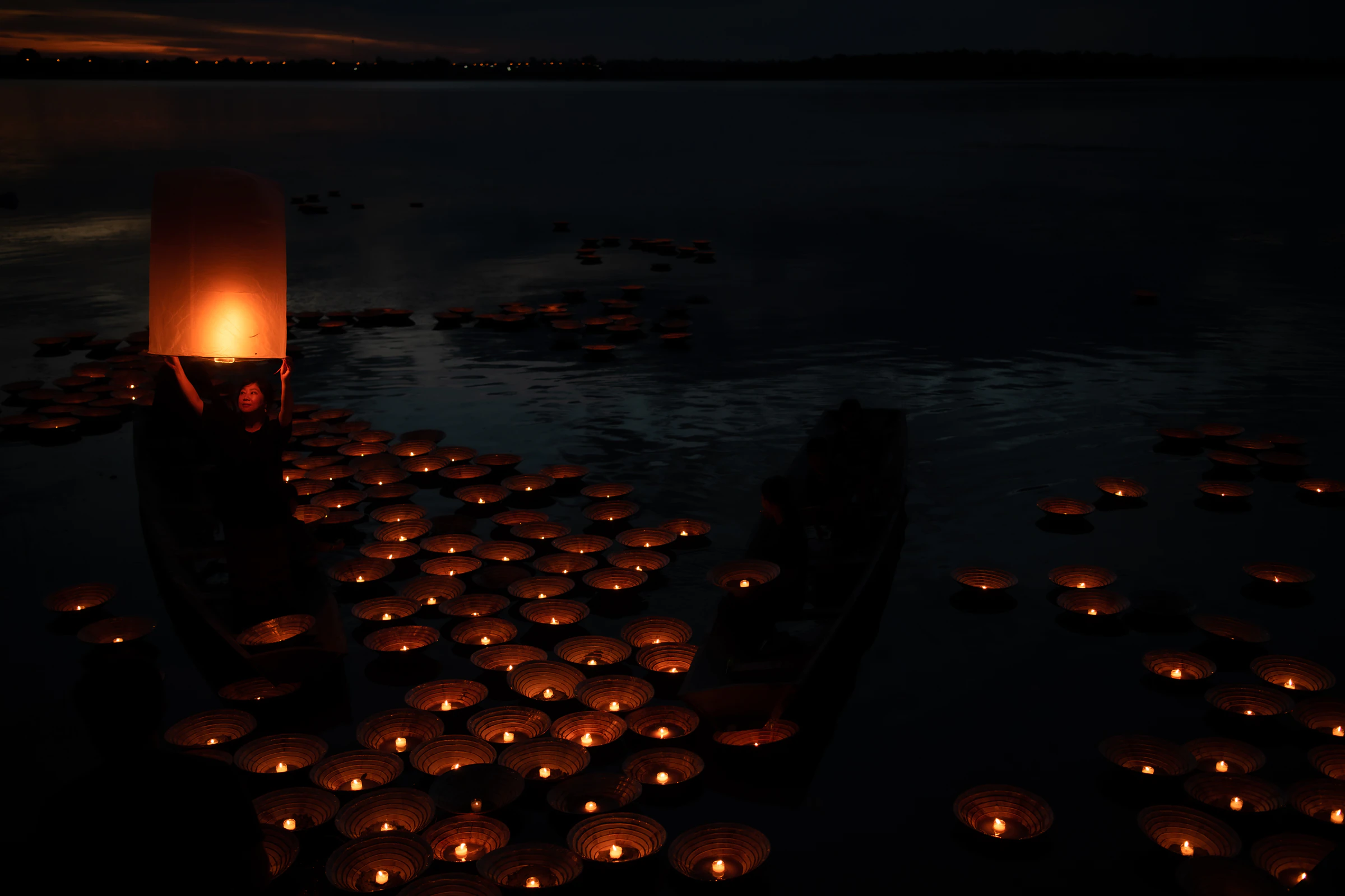 Candle-lit krathongs floating on dark water at night, with a glowing lantern above