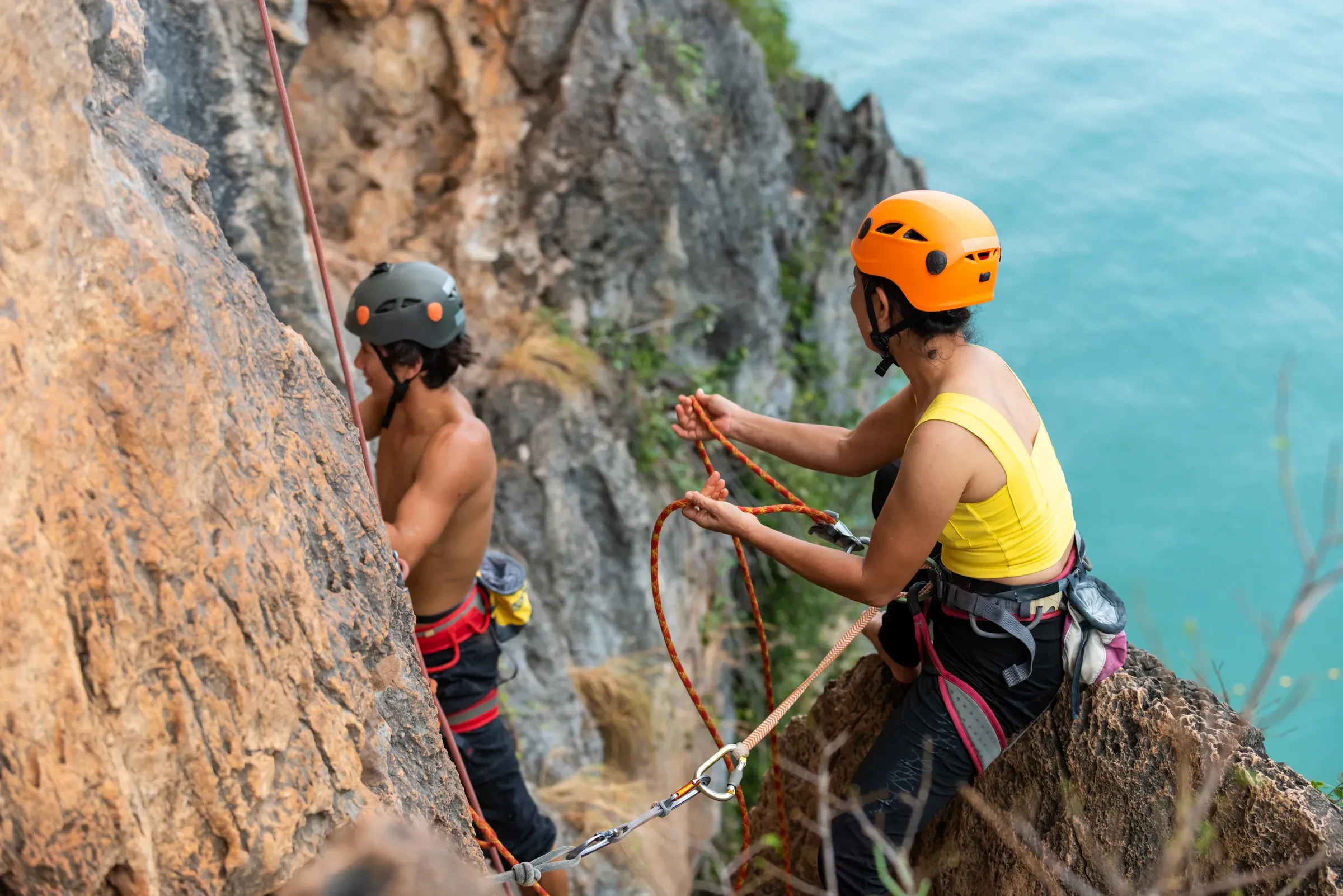 Two climbers in helmets handling a rope on limestone above turquoise water