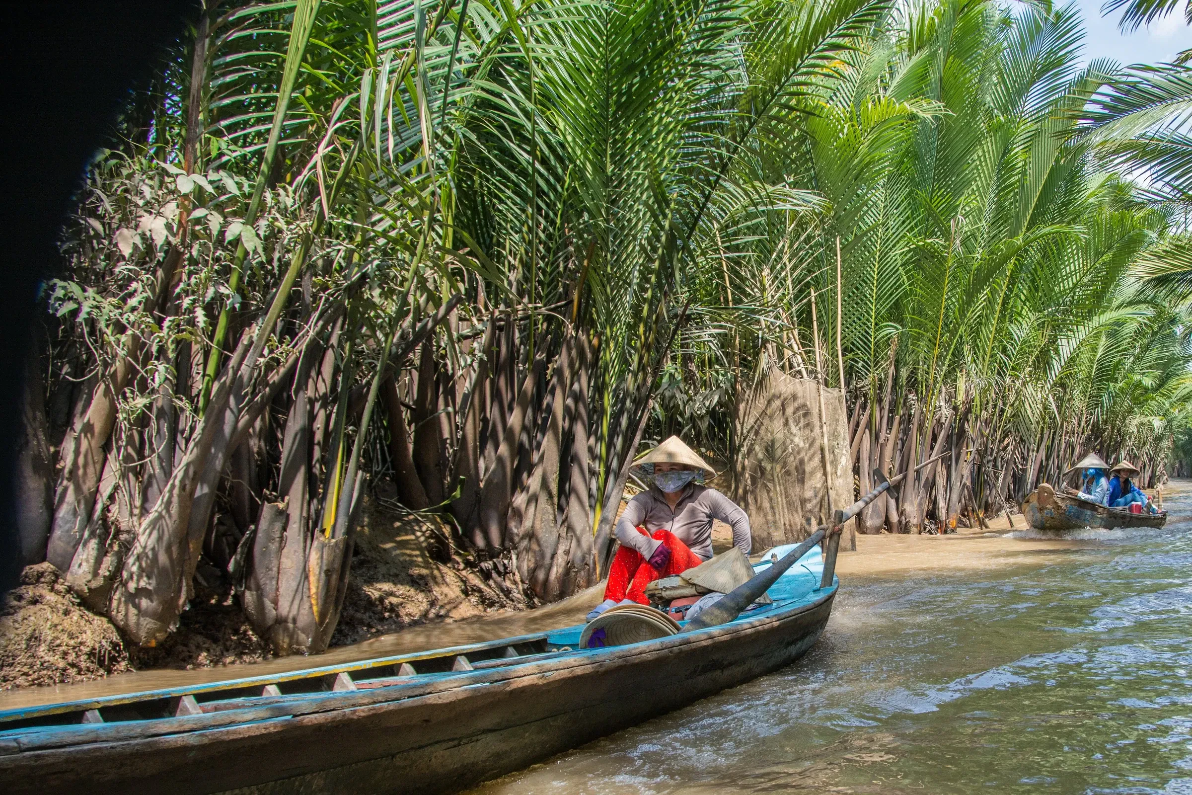 A rower in a conical hat on a palm-lined canal in the Mekong Delta