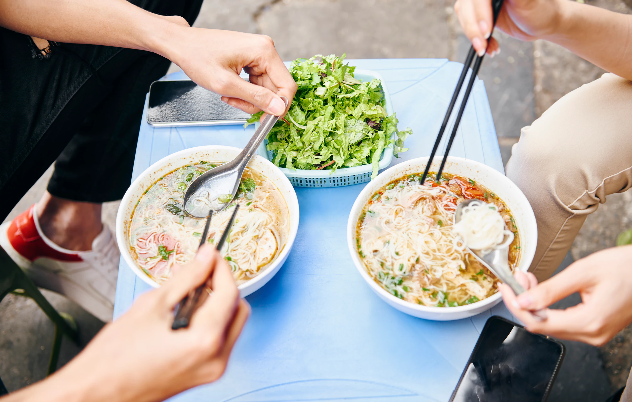 Two people sharing bowls of noodle soup at a street food table