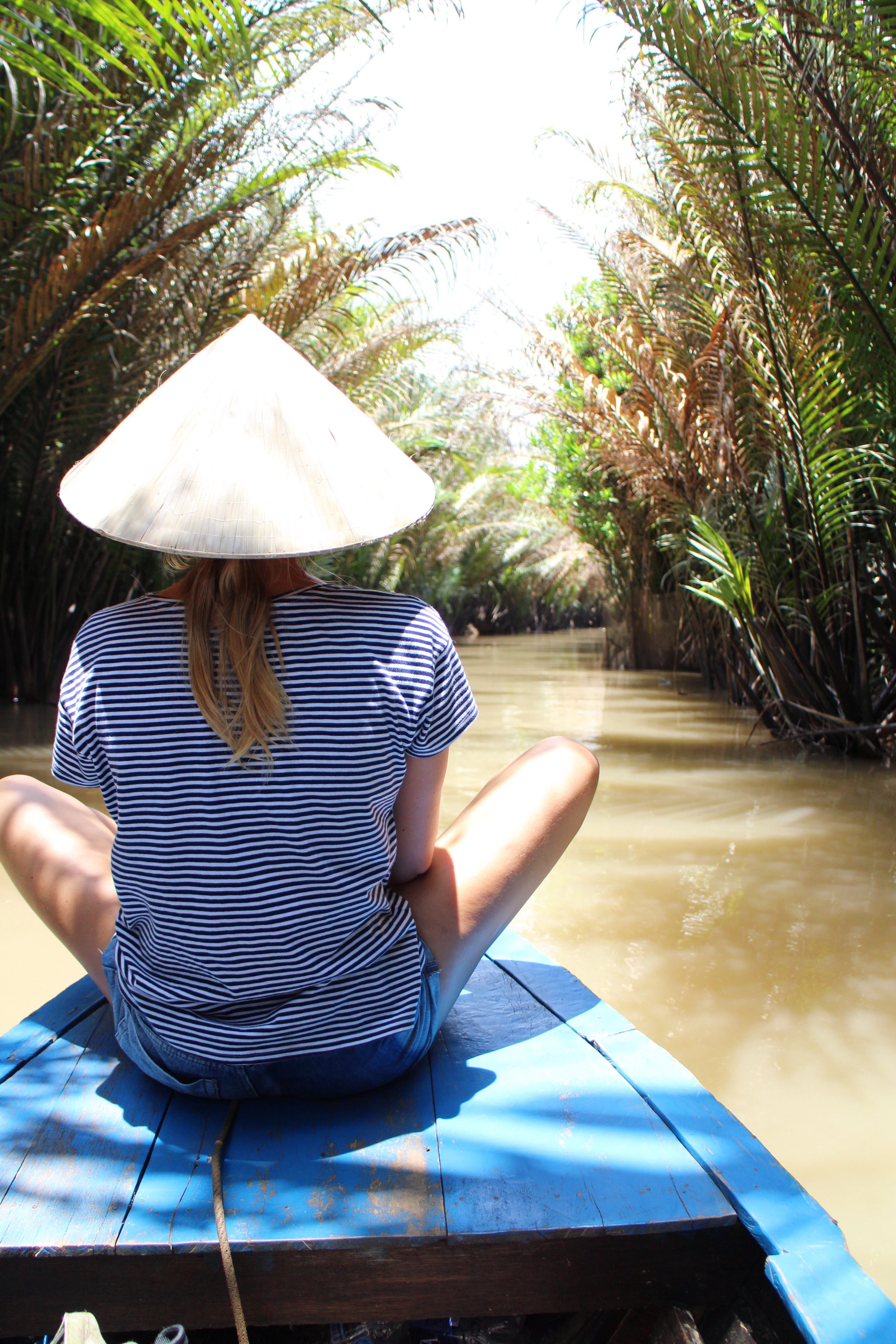 A traveller in a conical hat on a boat in a narrow canal lined with water coconut palms